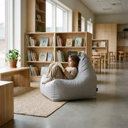 A cozy, minimalist children's library with a child reading in a grey beanbag chair, wooden bookshelves, and natural light.
