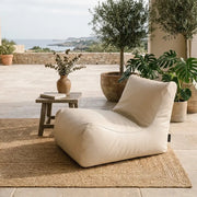 Sittsäck Lounge 100 utomhus beige bredvid ett litet bord på en terrass med växter och havsutsikt.