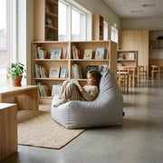 A cozy, minimalist children's library with a child reading in a grey beanbag chair, wooden bookshelves, and natural light.