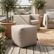 Modern outdoor patio with a beige fabric ottoman, comfortable armchairs, potted olive trees, and a natural jute rug.