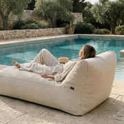 A woman relaxes on a modern, beige outdoor beanbag lounge chair by a serene swimming pool in a Mediterranean garden.