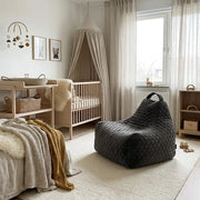 Cozy neutral nursery with a wooden crib, canopy, changing table, and a dark grey quilted beanbag chair on a soft rug.