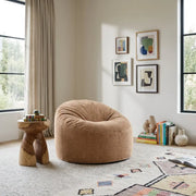 Modern living room corner featuring a tan corduroy bean bag chair, wooden side table, colorful art prints, and patterned rug.
