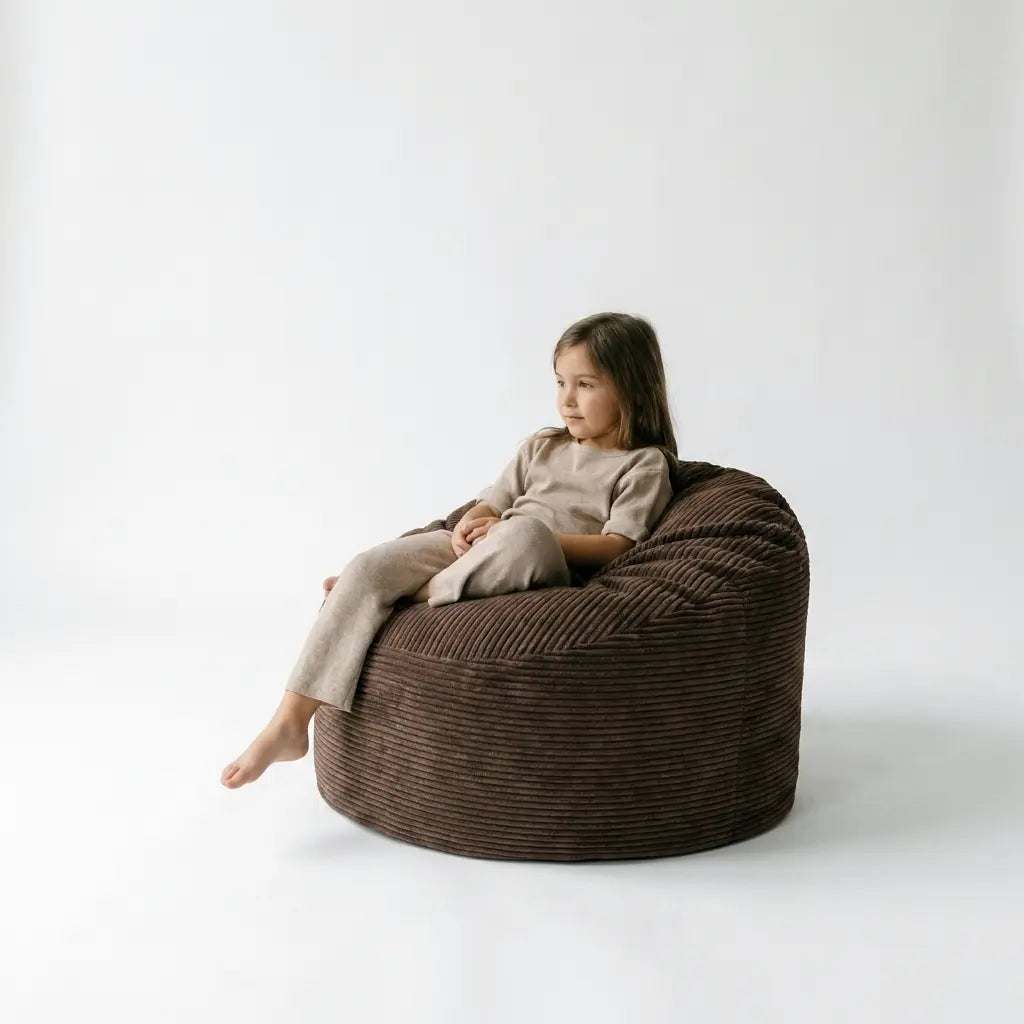 A young girl sits comfortably in a chocolate brown corduroy Barnfåtölj Sittsäck Om 85 Waves beanbag chair against a white wall.